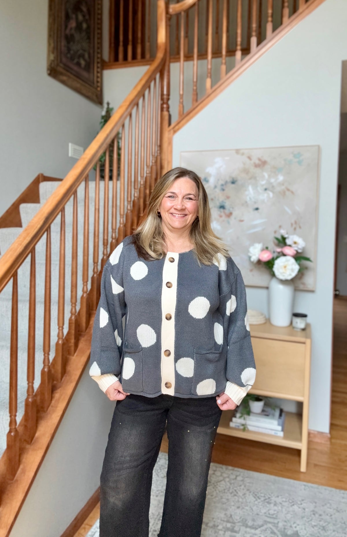 A woman stands in a home interior in front of a wooden staircase, wearing a gray cardigan with large white polka dots and cream trim, paired with dark jeans.
