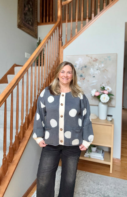 A woman stands in a home interior in front of a wooden staircase, wearing a gray cardigan with large white polka dots and cream trim, paired with dark jeans.