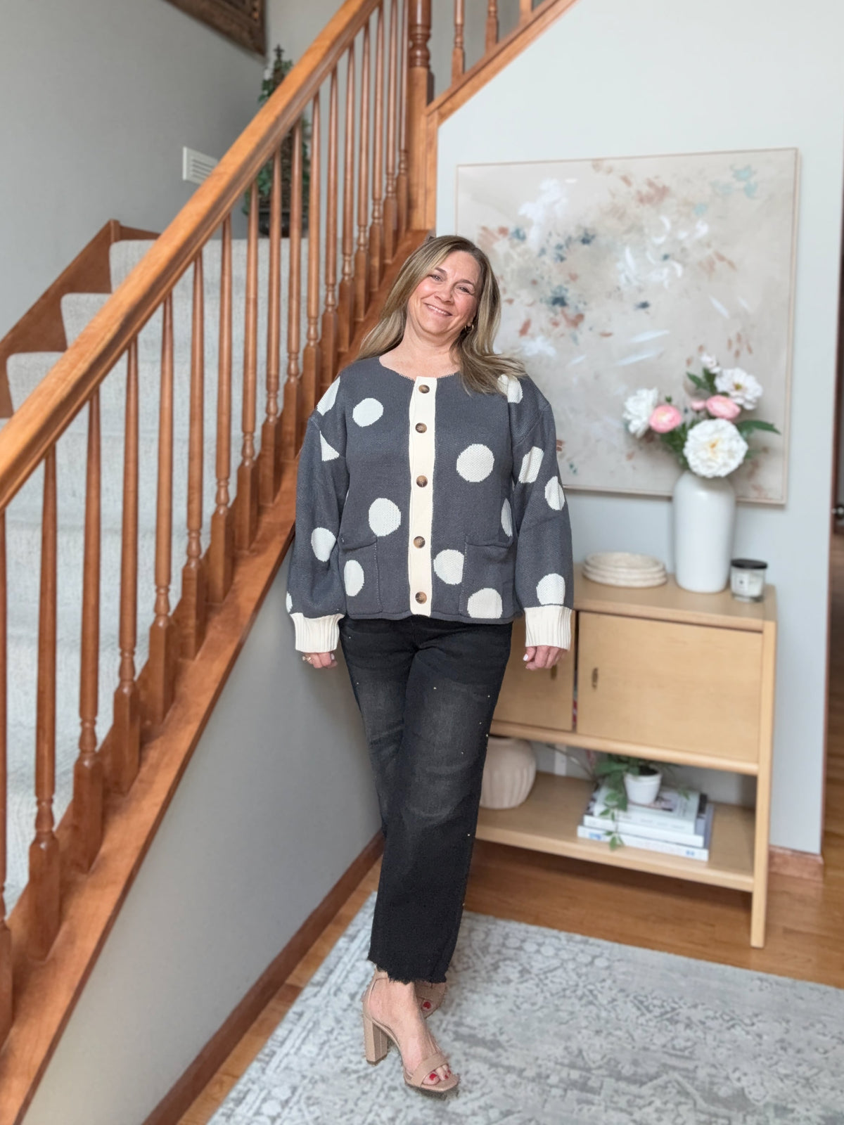A woman stands in a home interior next to a wooden staircase, wearing a dark gray cardigan with large white polka dots and cream buttons, paired with black jeans and beige heels.