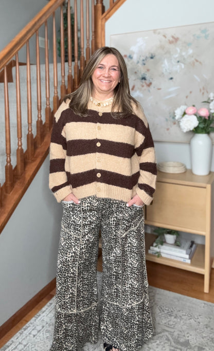 A woman stands indoors wearing a brown and beige striped knit cardigan with buttons, paired with wide-leg patterned pants. The setting includes a wooden staircase, a light wood side table with flowers, and a neutral wall.