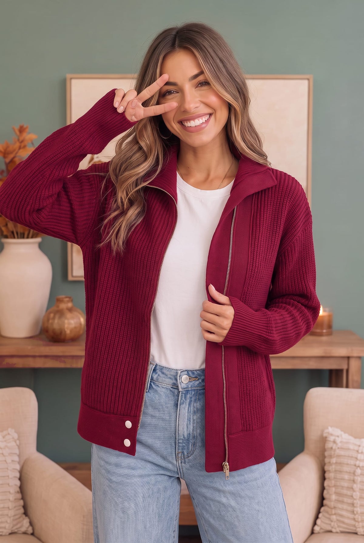 A woman wearing a burgundy ribbed zip-up cardigan over a white t-shirt and light blue jeans, posing in a cozy living room setting.