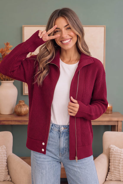 A woman wearing a burgundy ribbed zip-up cardigan over a white t-shirt and light blue jeans, posing in a cozy living room setting.