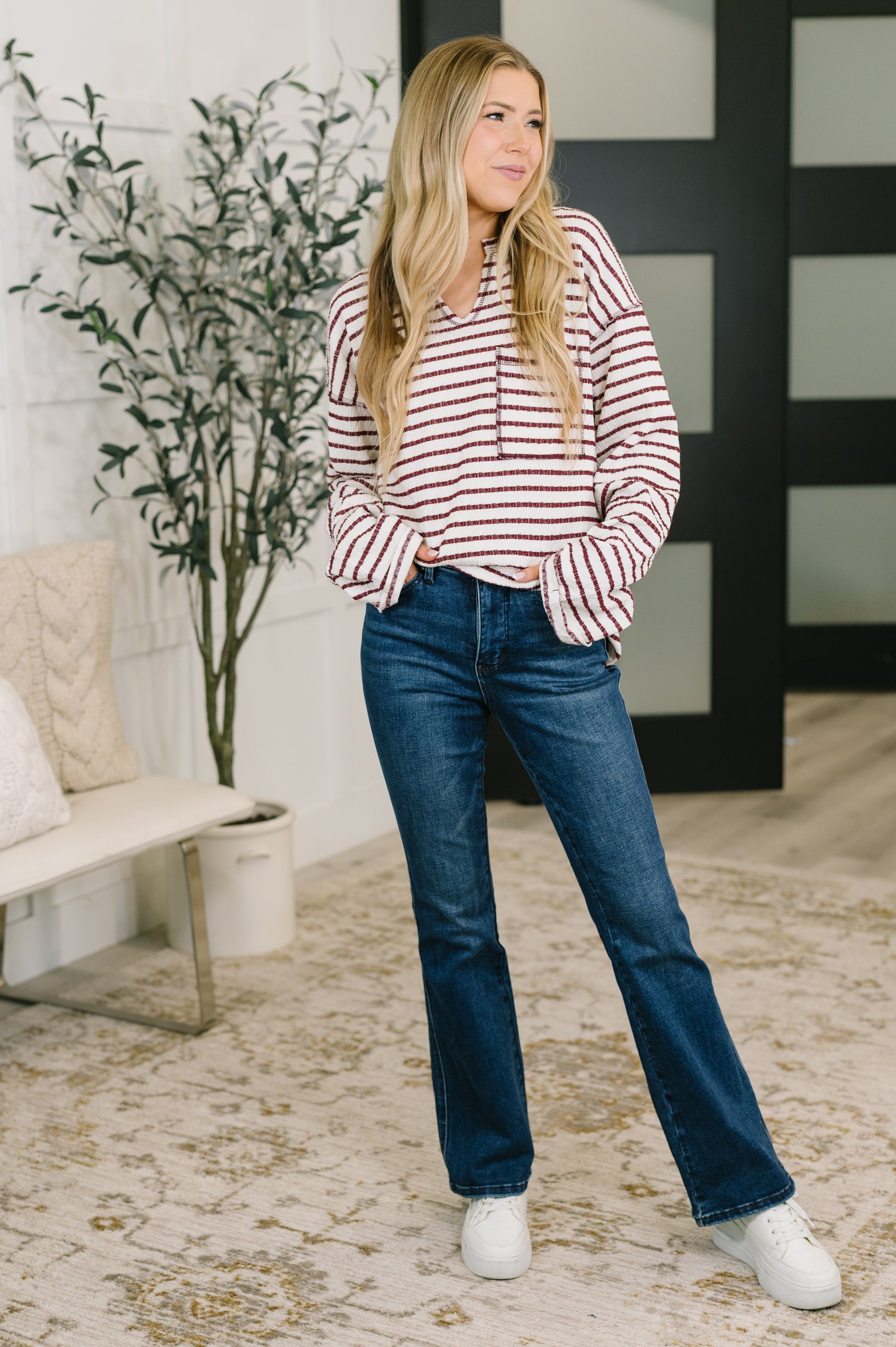 A woman modeling a red and white striped long-sleeve top with blue flare jeans and white sneakers, standing in a modern indoor room with a bench, potted plant, and patterned rug.