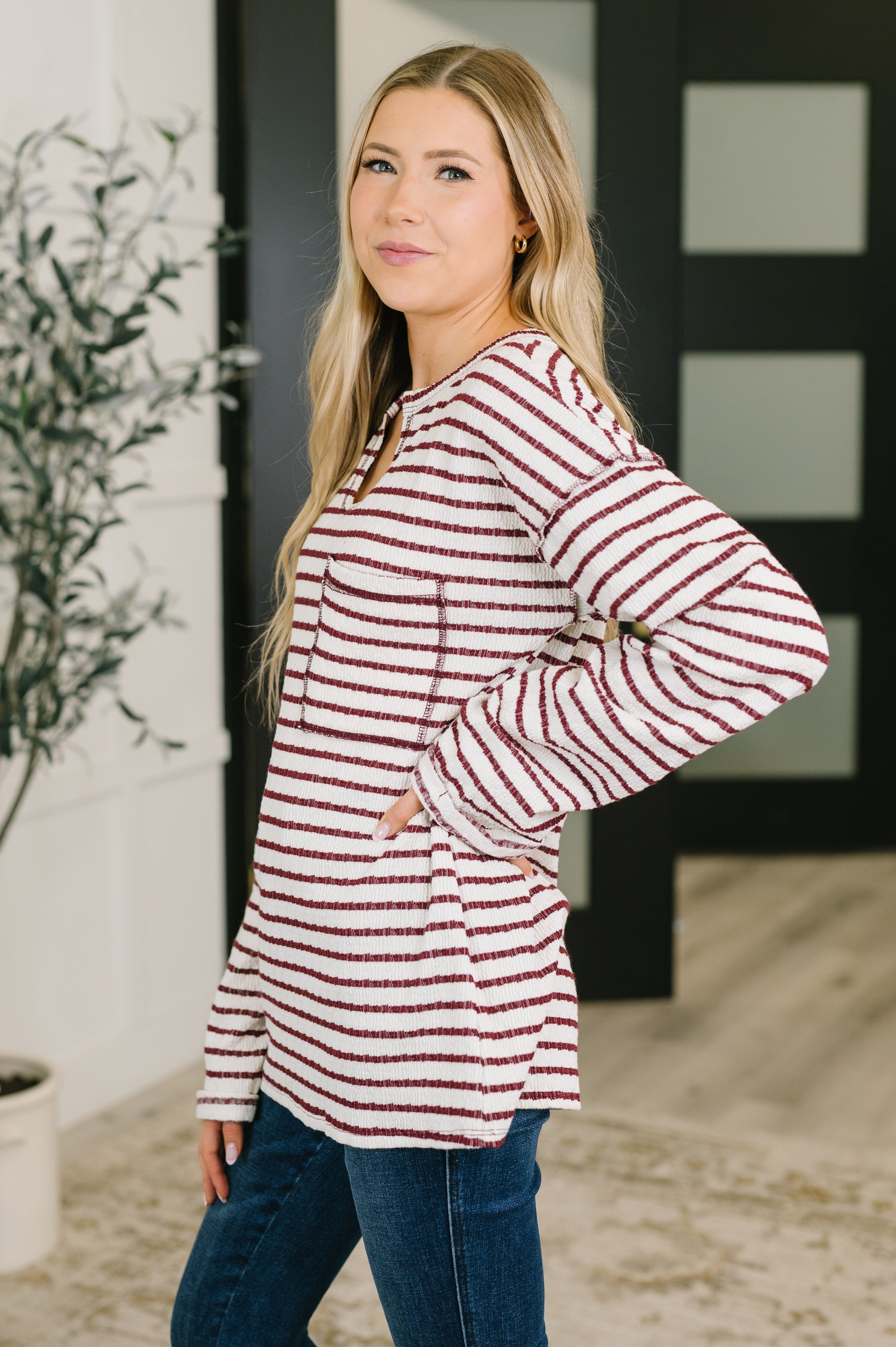 A woman modeling a long-sleeve knit top with horizontal red and white stripes, paired with blue jeans, standing in a modern indoor setting.