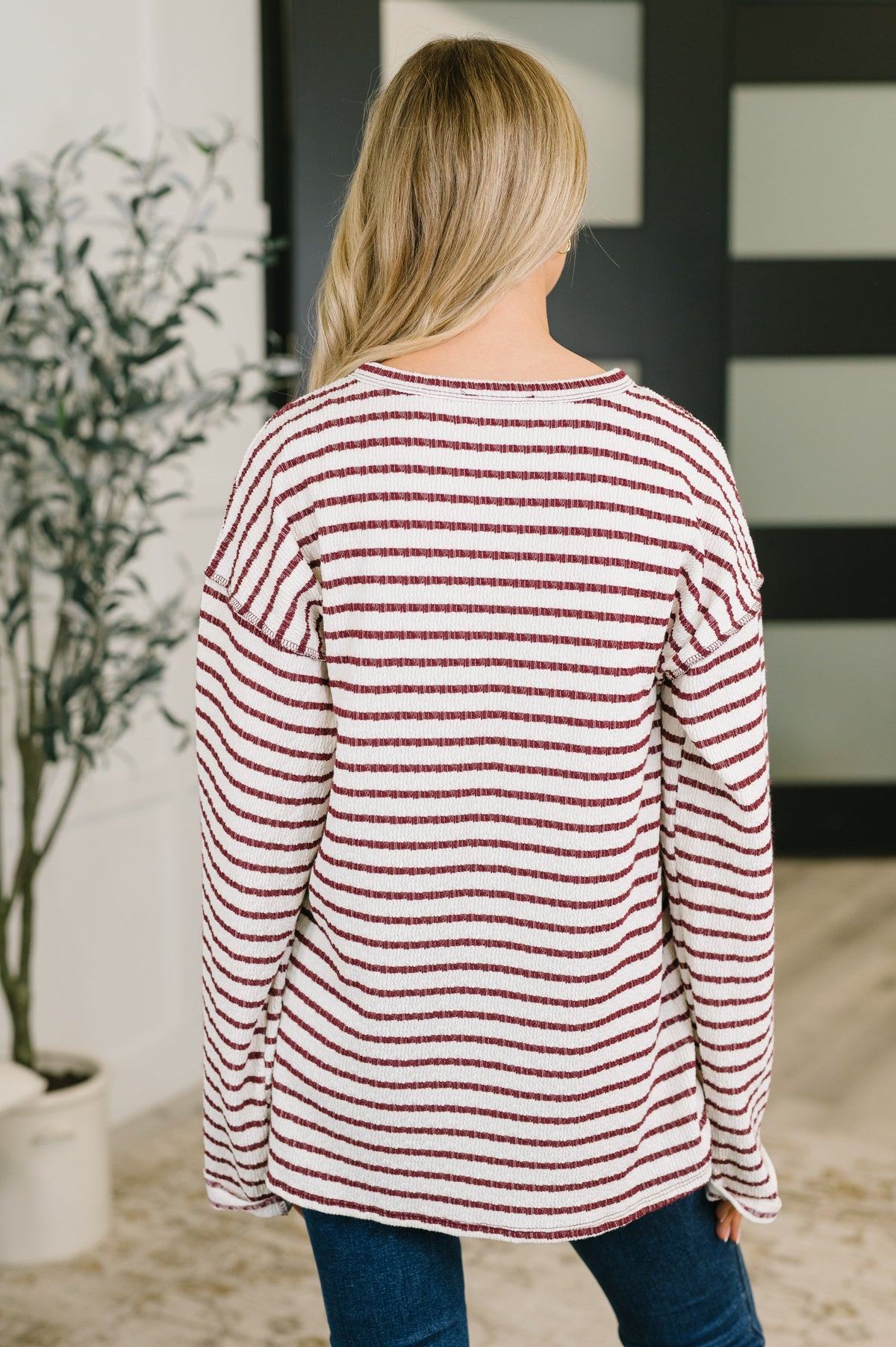 A woman viewed from behind wearing a long-sleeve knit top with thin horizontal red and off-white stripes, paired with blue jeans in an indoor setting.