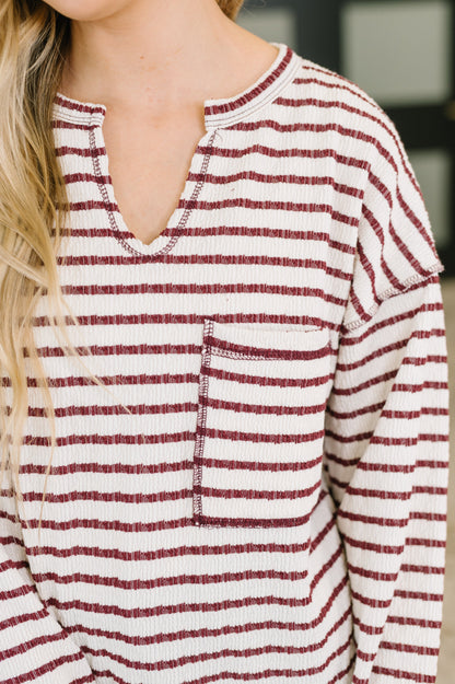 A close-up of a woman wearing a long-sleeve knit top with horizontal burgundy and off-white stripes, featuring a split neckline and a chest pocket.
