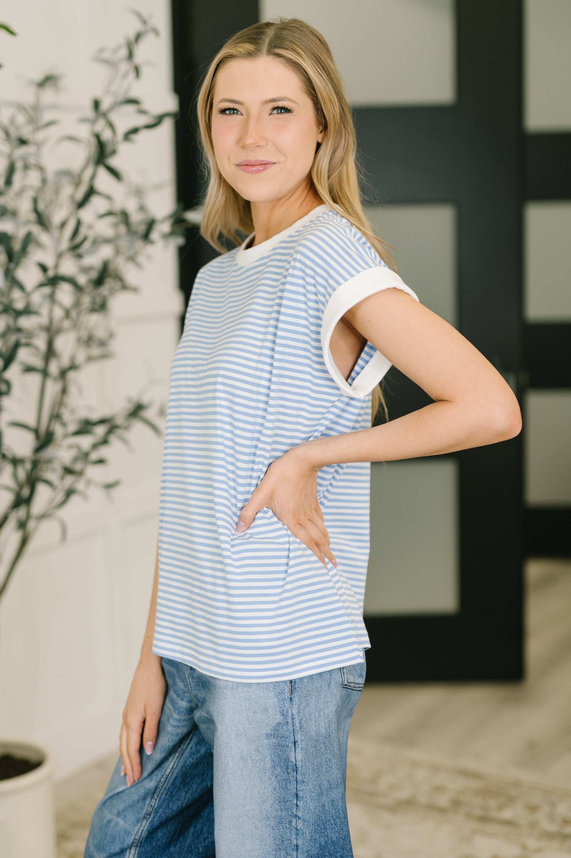 Woman wearing a striped shirt and jeans standing indoors with a plant in the background