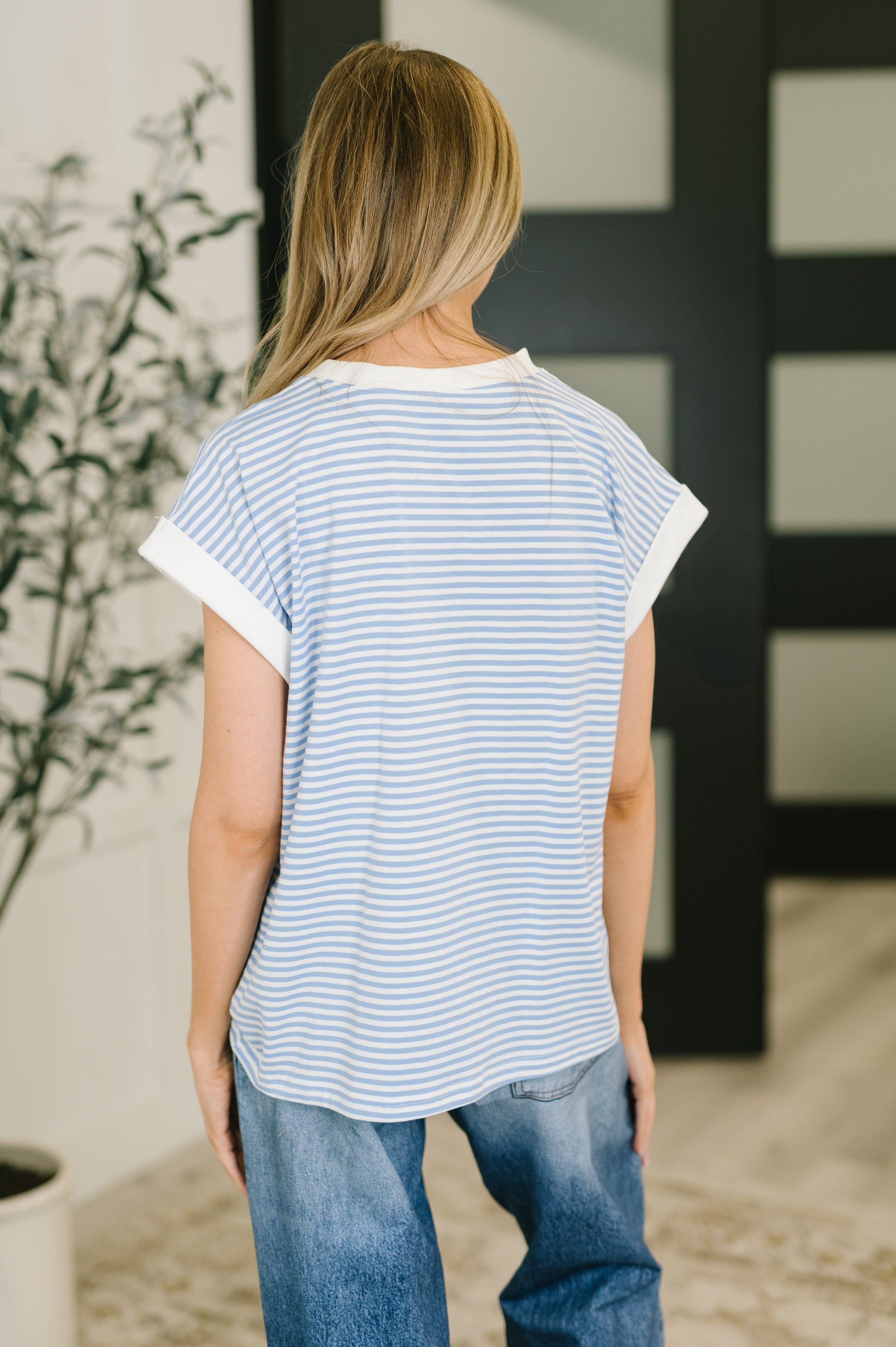 Person wearing a striped shirt and jeans standing in a room with a plant and decorative wall.