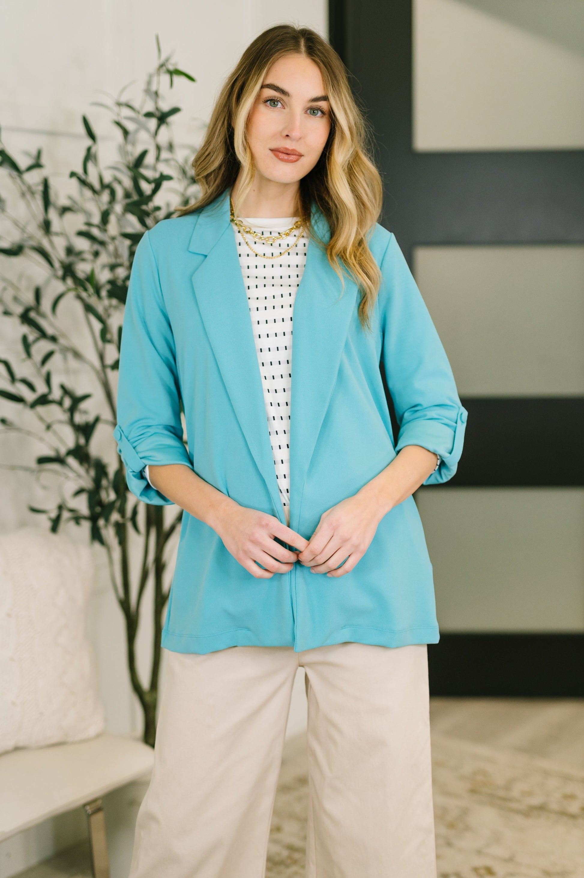 Woman wearing a light blue jacket over a patterned top and beige pants, standing indoors.