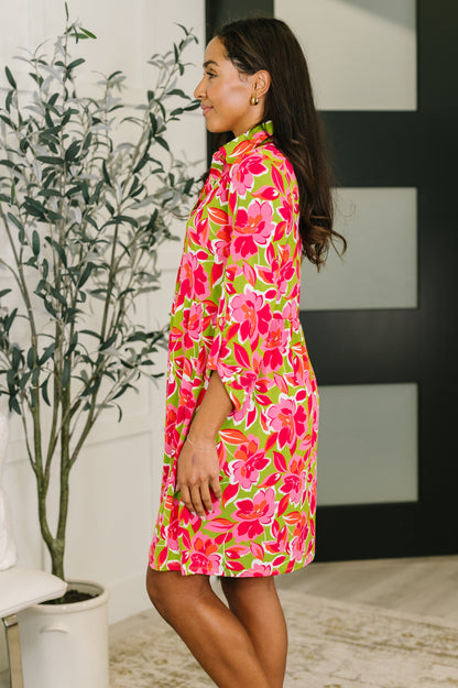 Woman wearing a colorful floral dress standing indoors with a plant in the background