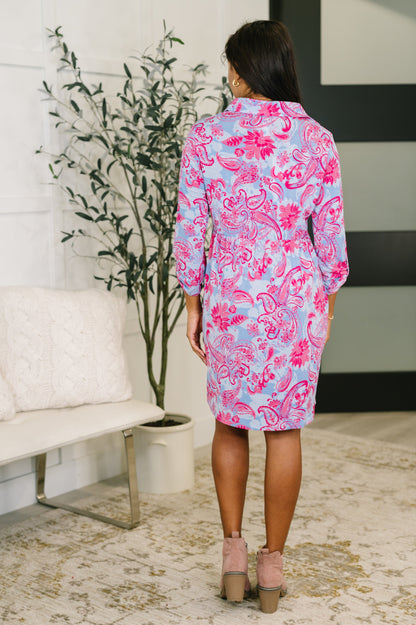Woman wearing a pink and blue patterned dress in a room with a plant and bench.