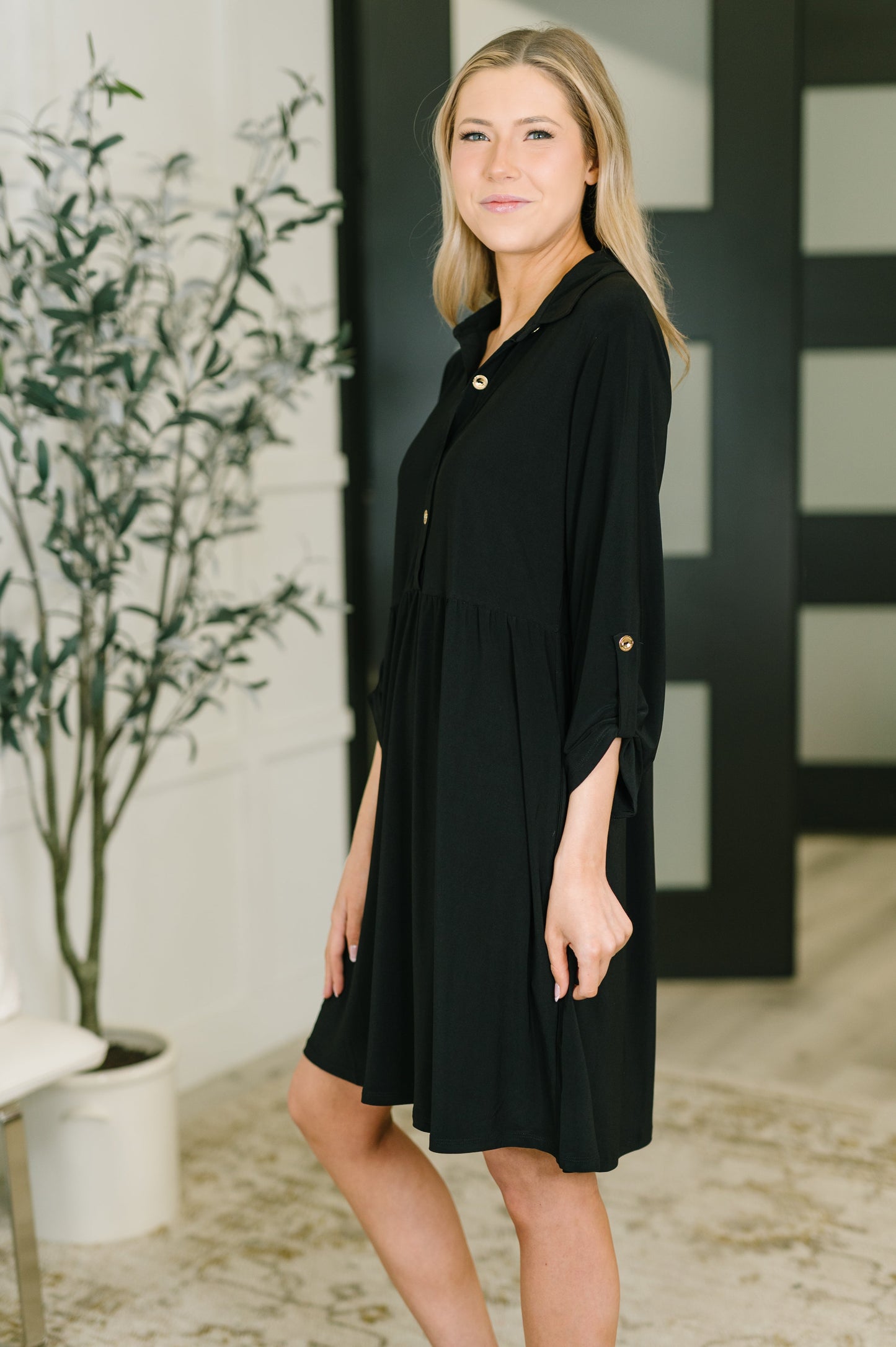 Woman wearing a black dress standing indoors with a plant and decorative wall in the background