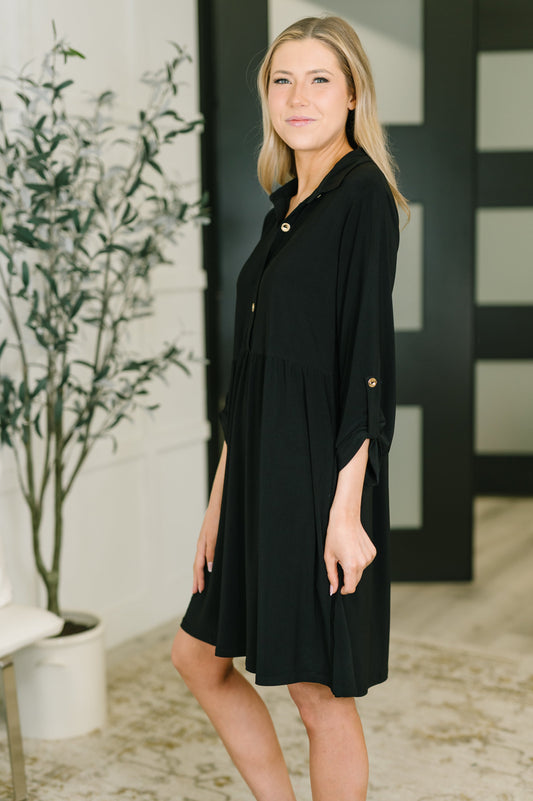 Woman wearing a black dress standing indoors with a plant and decorative wall in the background