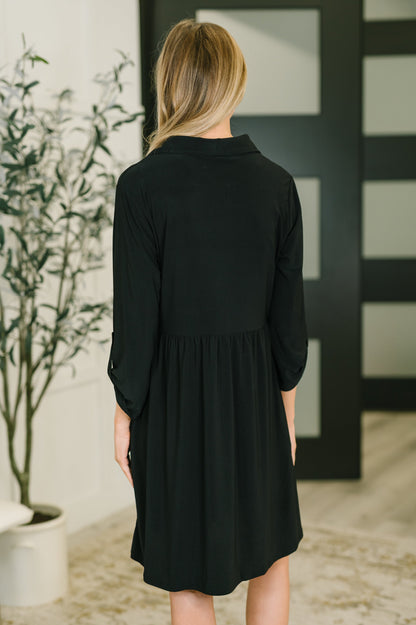 Woman wearing a black dress standing in a room with a plant and abstract art on the wall.