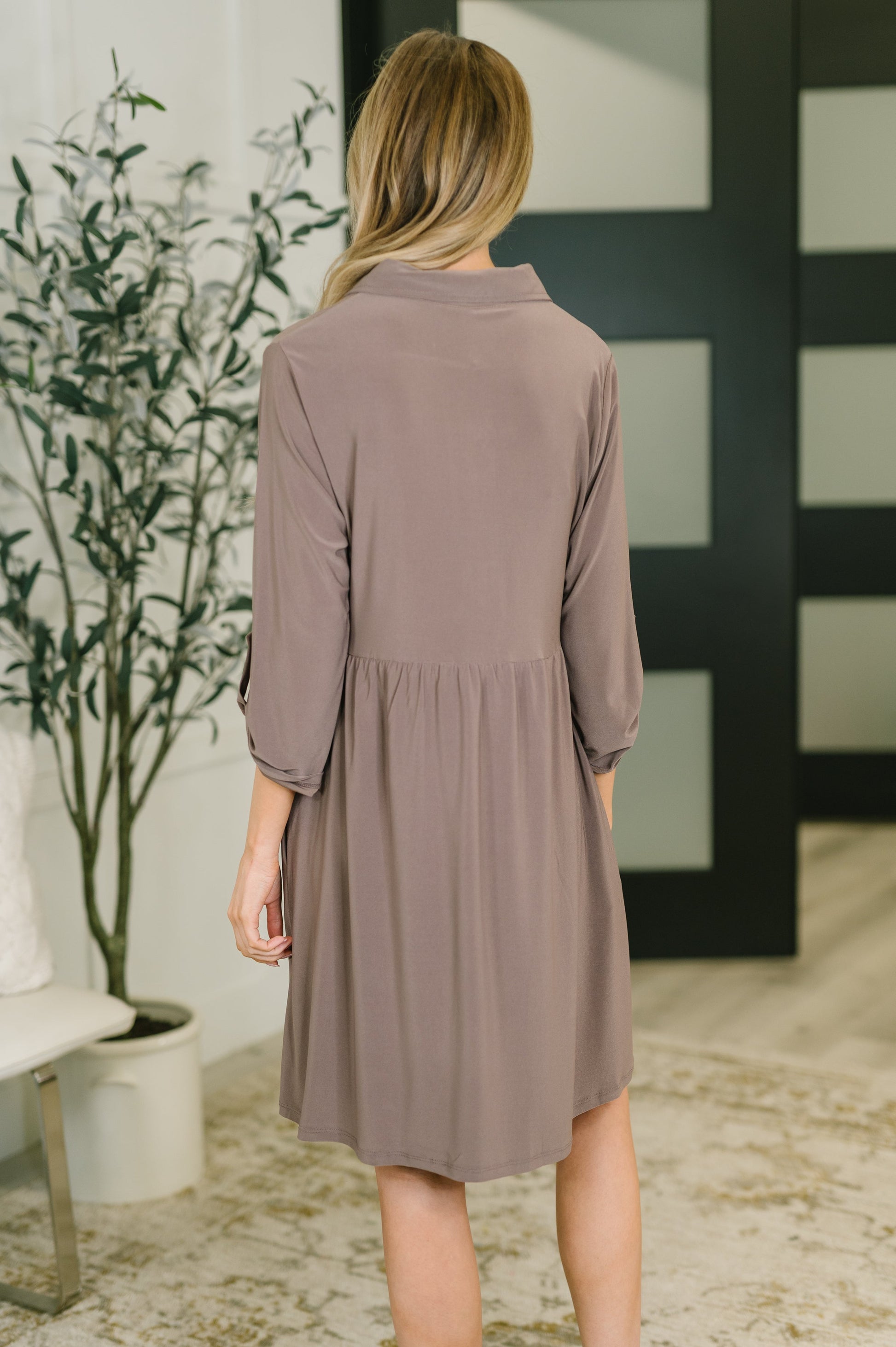 Woman wearing a brown dress standing in a room with a plant and a bench.