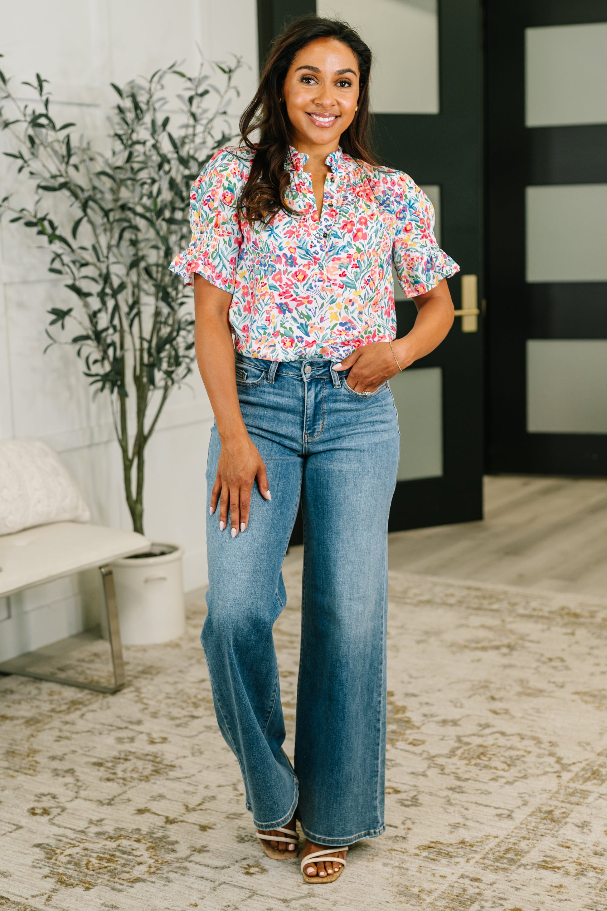 Woman wearing a floral blouse and blue jeans standing in a room with a plant and door in the background.