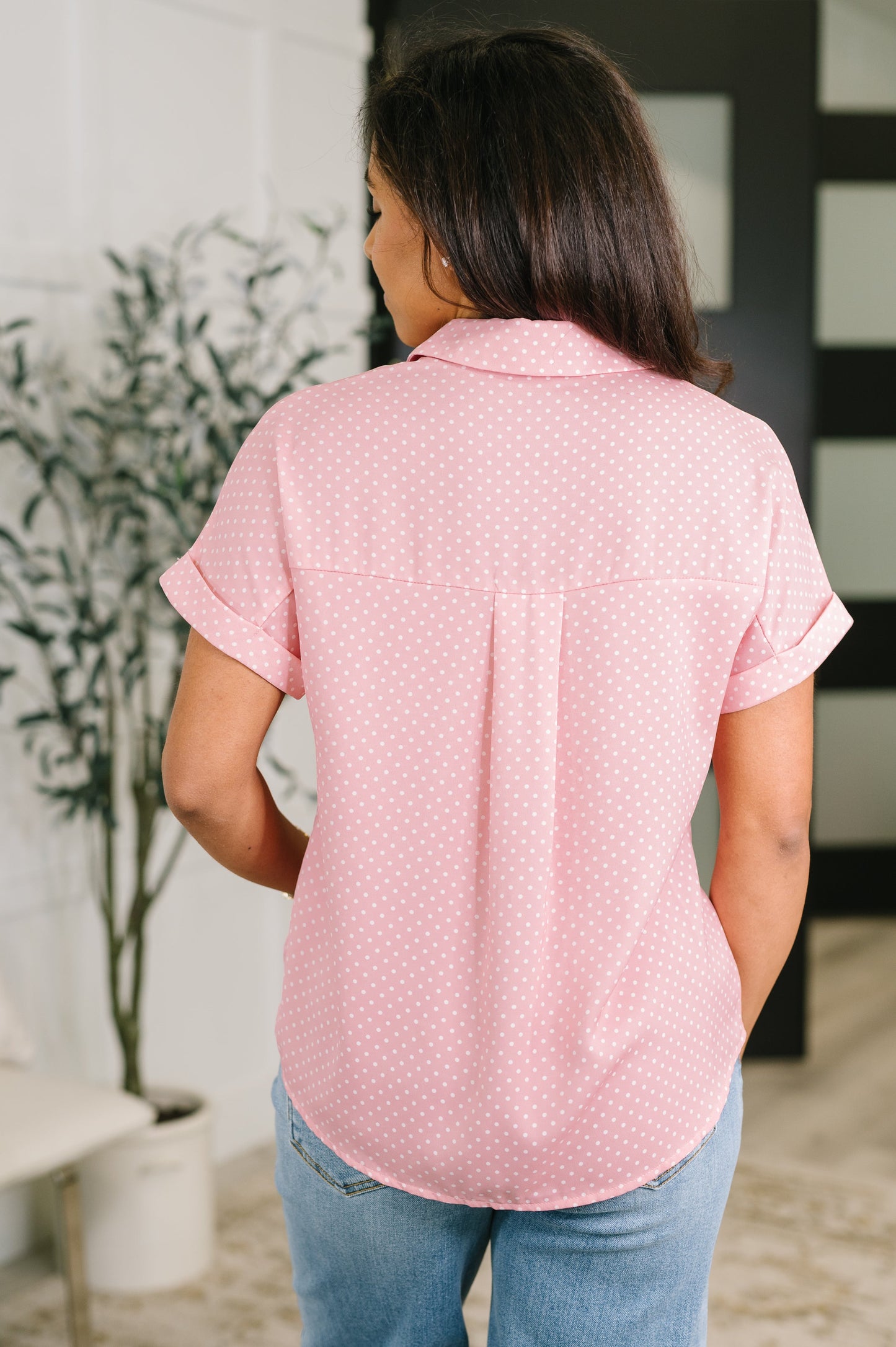 A woman viewed from behind wearing a light pink short-sleeve button-up shirt with a small white polka dot pattern and light blue jeans, standing in an indoor setting.