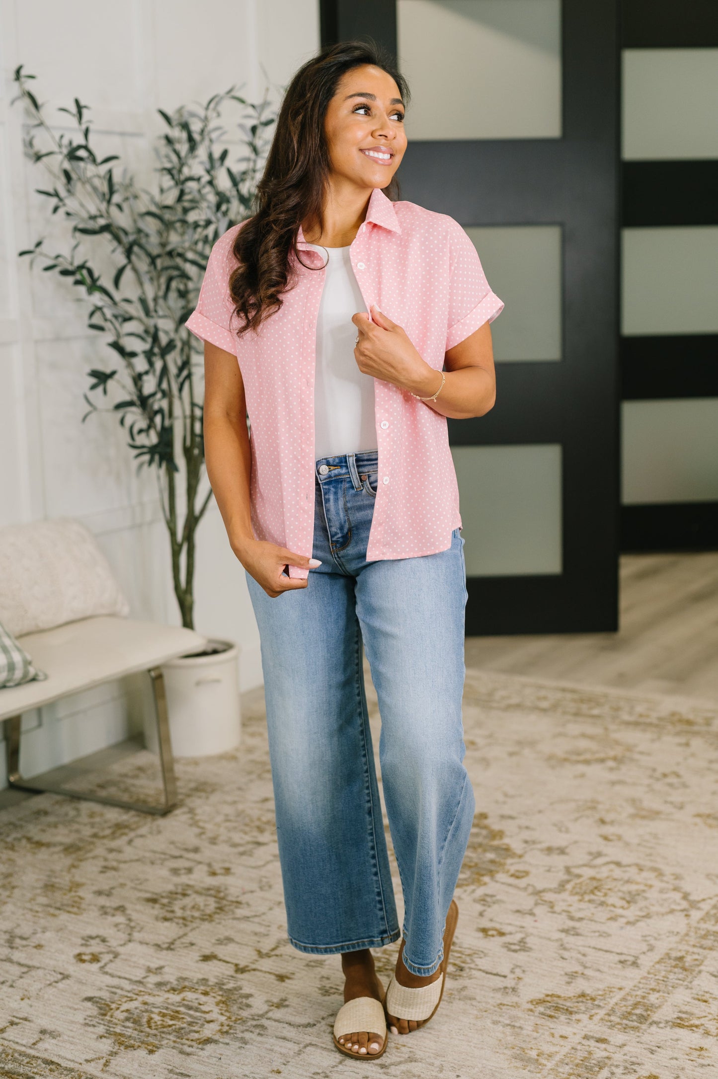 A woman modeling a light pink short-sleeve button-up shirt over a white top, paired with blue wide-leg jeans and brown sandals, standing in a modern indoor setting.