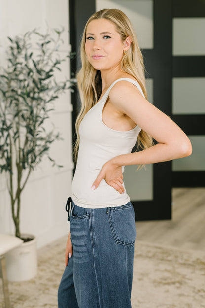 A woman modeling a fitted white ribbed tank top and blue jeans, standing indoors with a potted plant and modern door in the background.