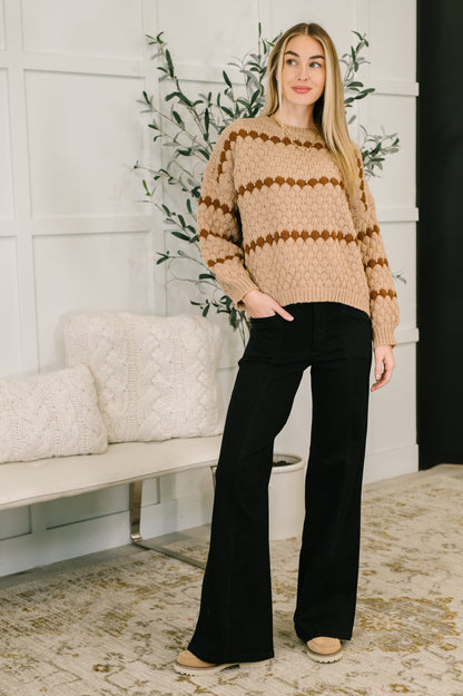 Woman wearing a beige and brown striped sweater with black pants in a living room setting.