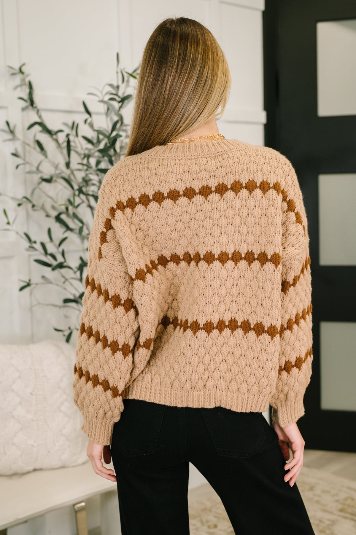 Person wearing a beige sweater with brown stripes in a room with a plant and white wall.