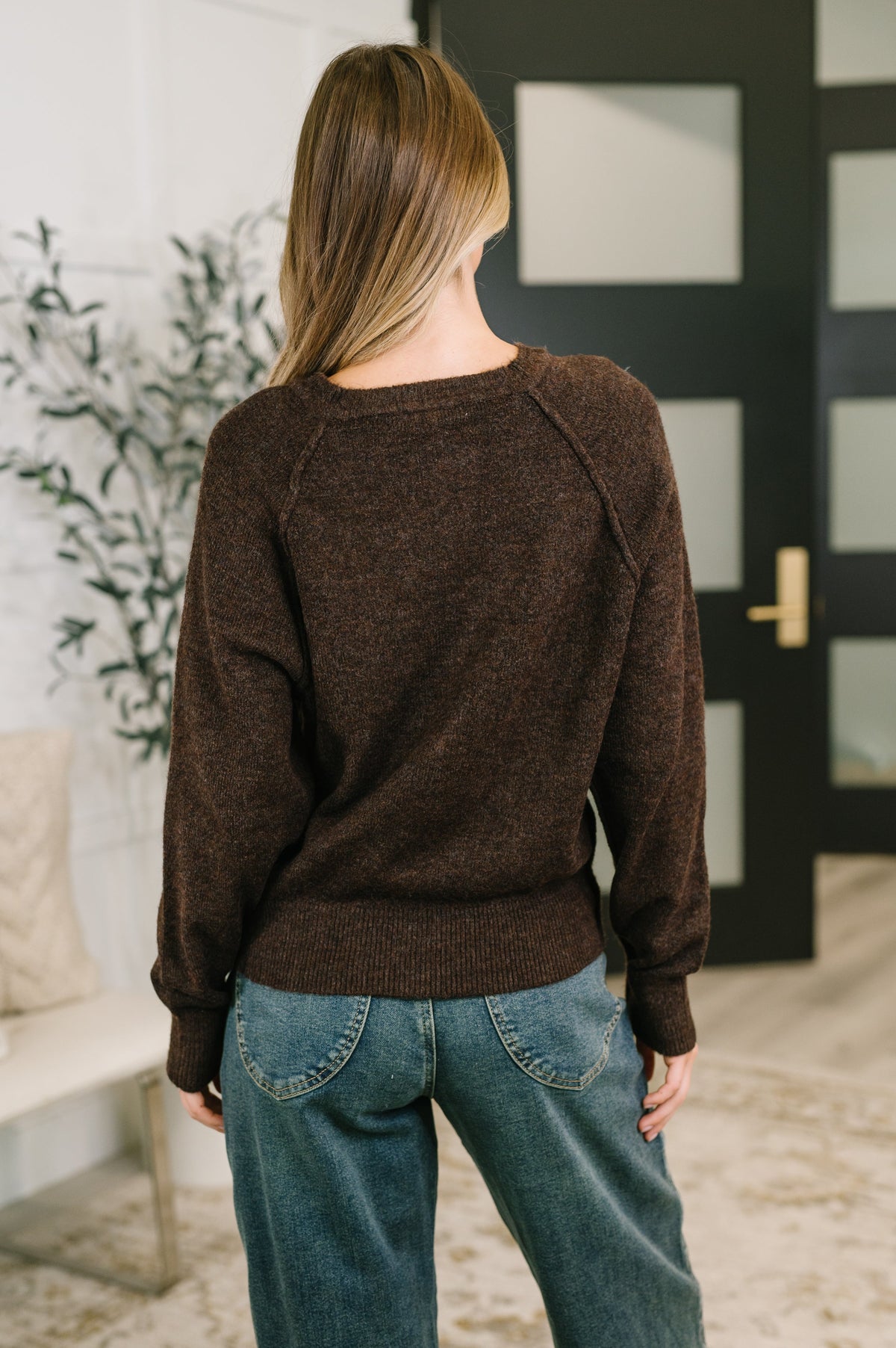 A woman viewed from behind wearing a dark brown knit sweater and blue jeans in an indoor setting with a black paneled door in the background.