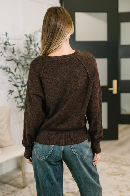 A woman viewed from behind wearing a dark brown knit sweater and blue jeans in an indoor setting with a black paneled door in the background.