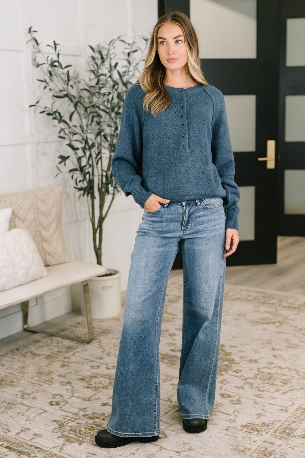 A woman stands indoors wearing a dusty blue knit long-sleeve henley top and wide-leg blue jeans, with her hands in her pockets. The setting includes a patterned rug, a bench with pillows, and a potted plant.