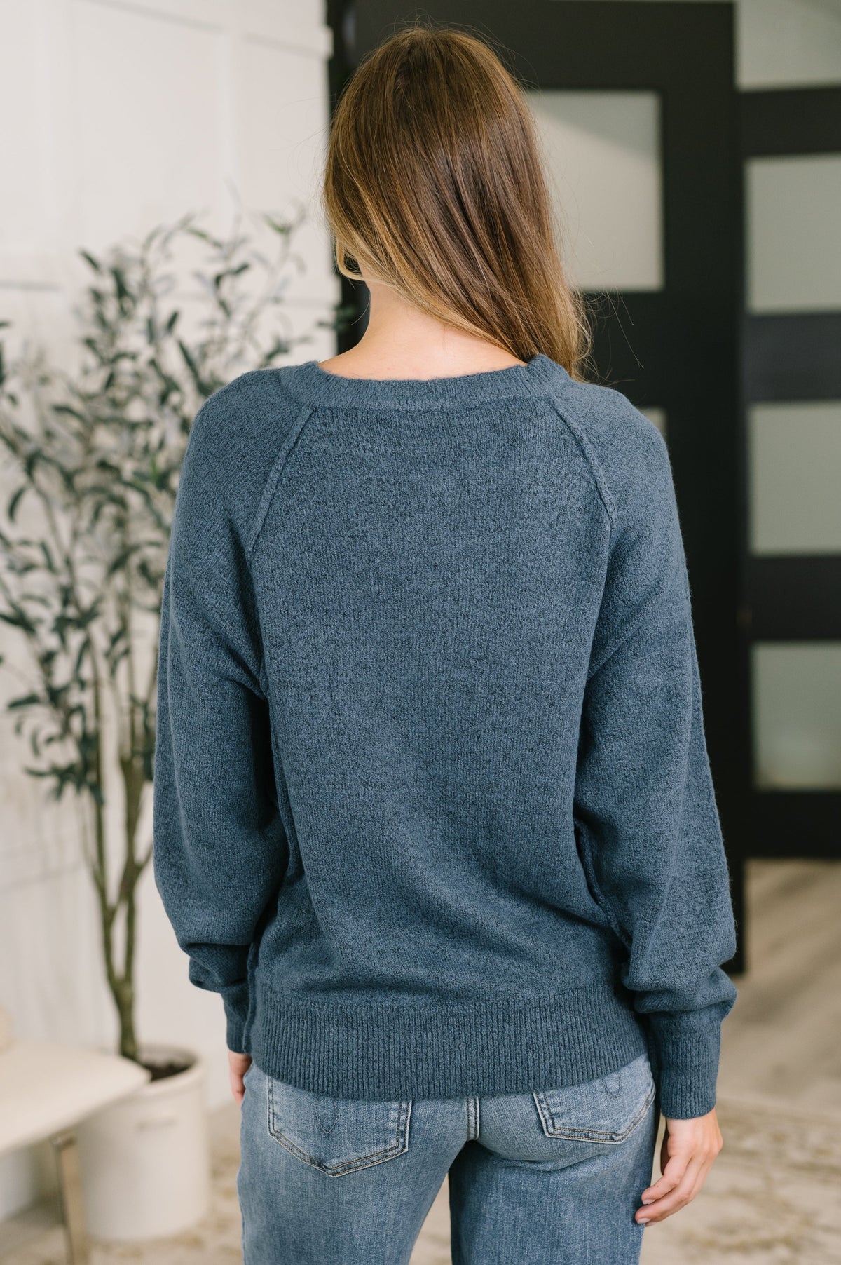 A woman viewed from behind wearing a heathered slate blue knit sweater and light blue jeans in an indoor setting.