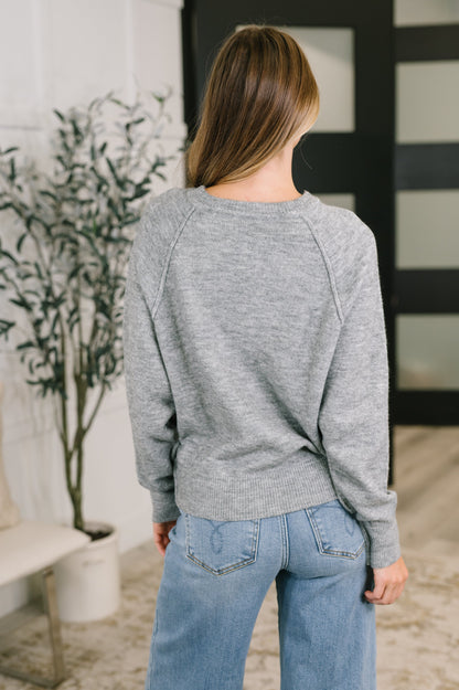 A woman viewed from behind wearing a heather gray long-sleeve crewneck sweater and light blue jeans in an indoor setting.