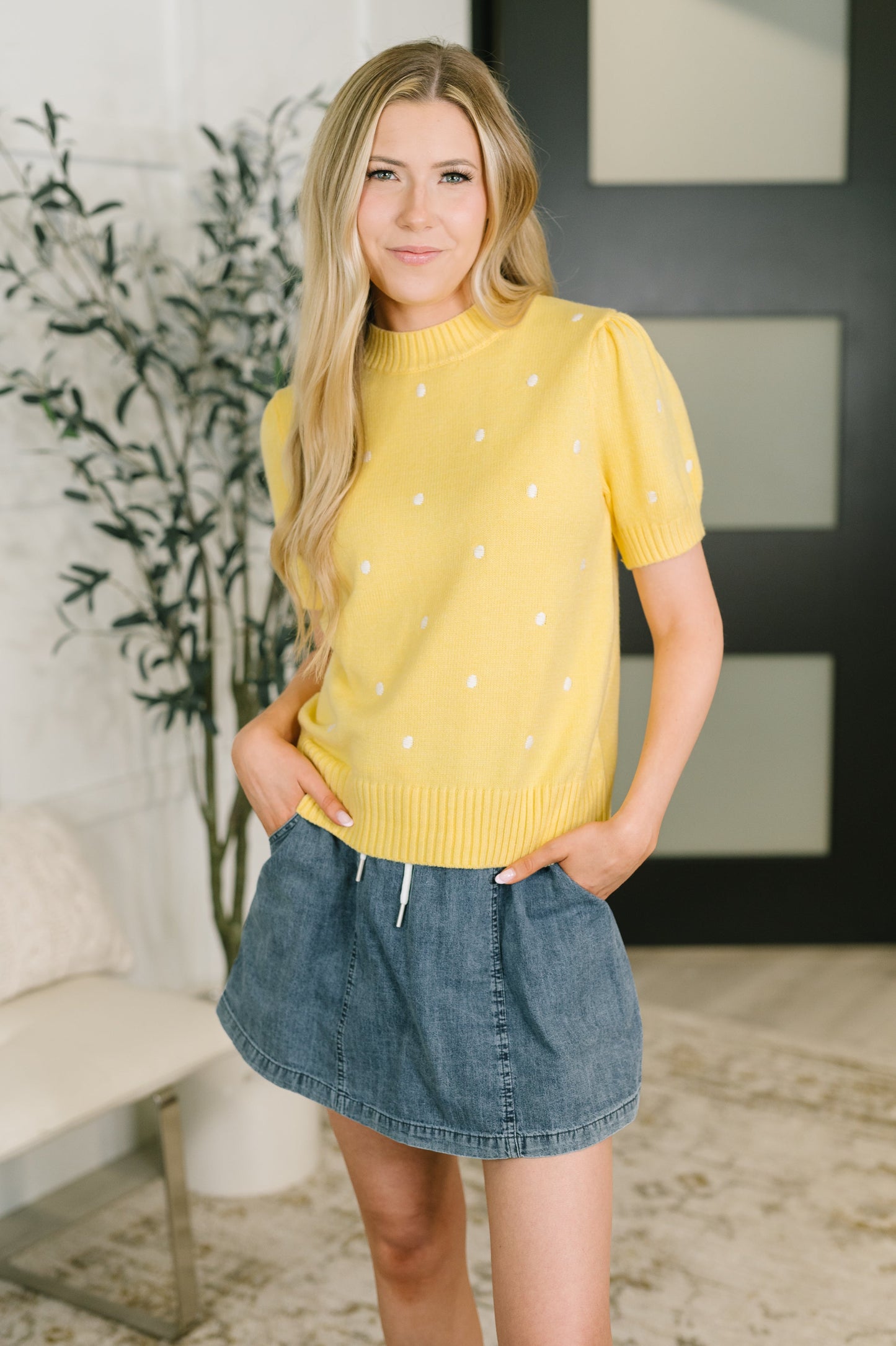 A woman modeling a bright yellow short-sleeve knit sweater with small white embroidered dots, paired with a denim skirt in an indoor setting.