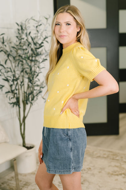 A woman modeling a bright yellow short-sleeve knit sweater with small white polka dots, paired with a denim mini skirt, standing in a modern indoor setting with a potted plant and dark-framed wall panels.