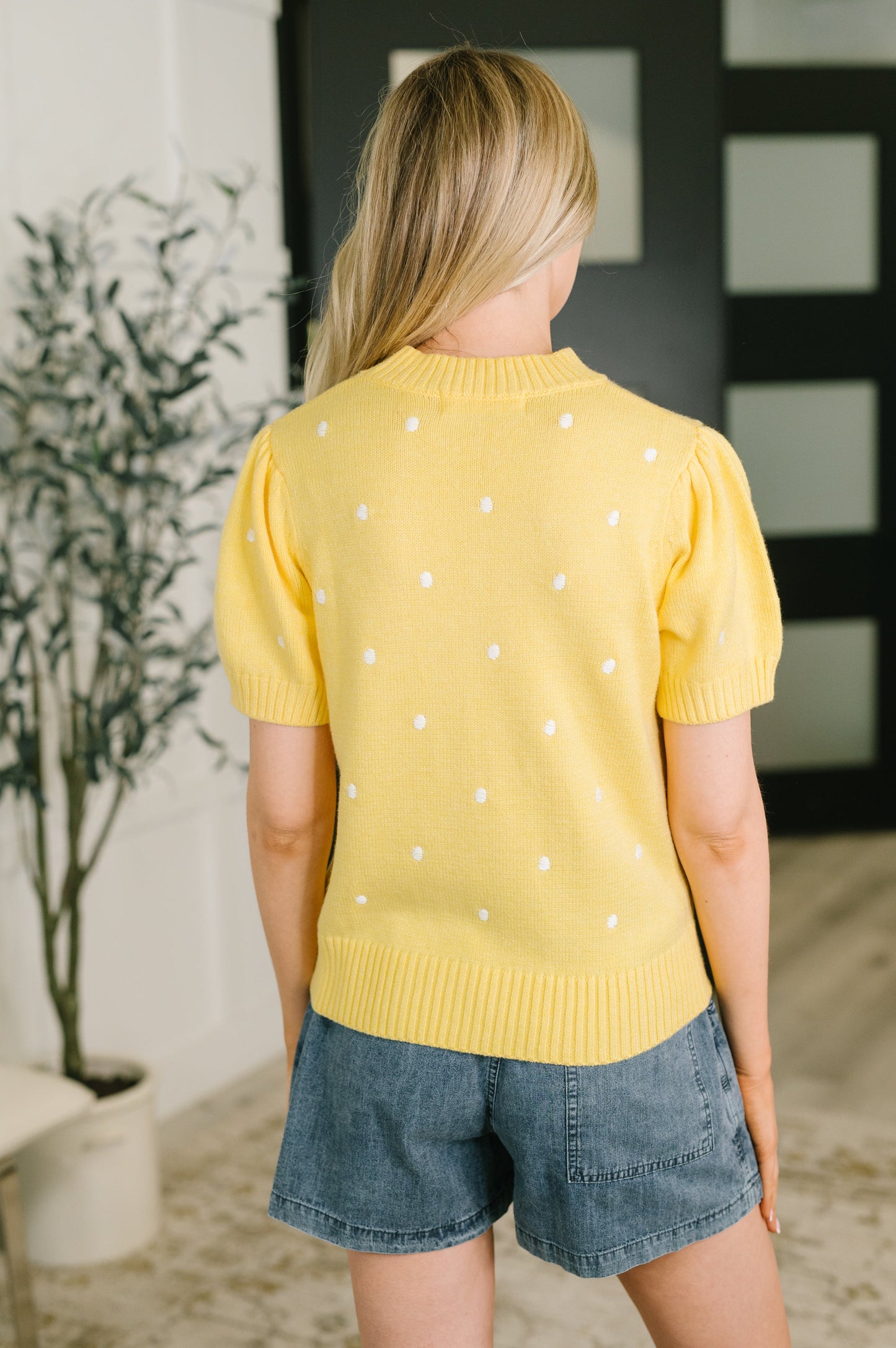 A woman viewed from behind wearing a yellow short-sleeve knit sweater with small white polka dots and blue denim shorts in an indoor setting.
