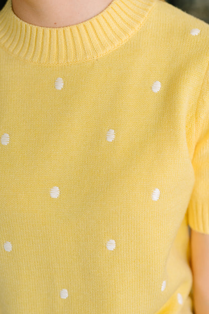 Close-up of a yellow knit top with small white polka dots, showing the ribbed crew neck and short sleeves.