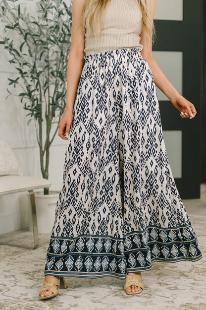 A woman wearing a cream knit top and high-waisted, wide-leg maxi pants with a navy blue and cream ikat pattern and a decorative border, standing in a modern indoor setting.