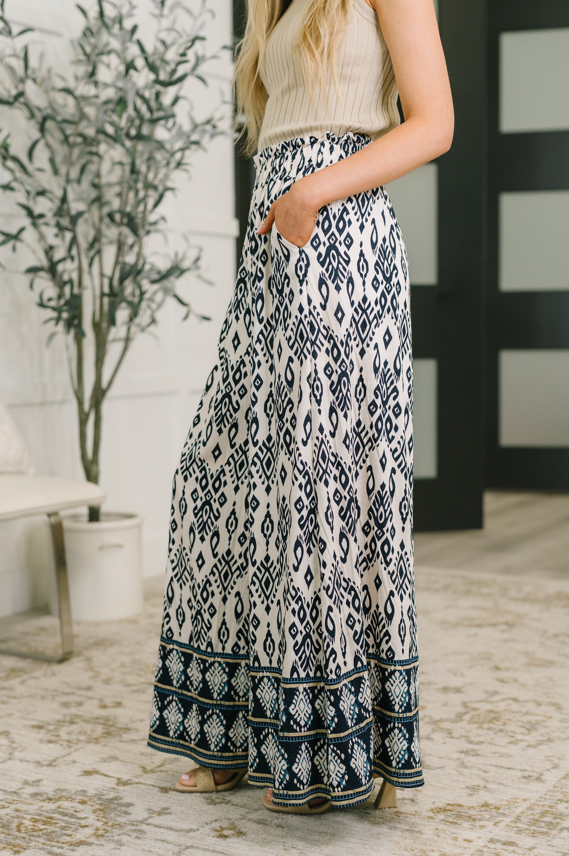 A woman wearing a cream ribbed knit sleeveless top and a long, wide-leg skirt with a navy and cream ikat pattern, standing in a modern indoor setting.