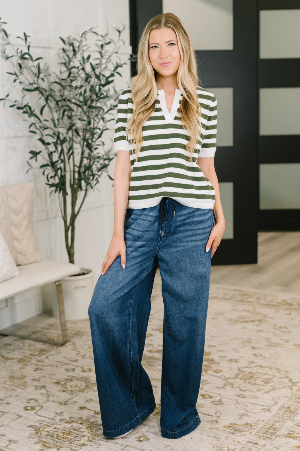 A woman modeling a short-sleeve green and white striped polo shirt with dark blue wide-leg jeans, standing in a modern indoor setting with a bench, potted plant, and patterned rug.
