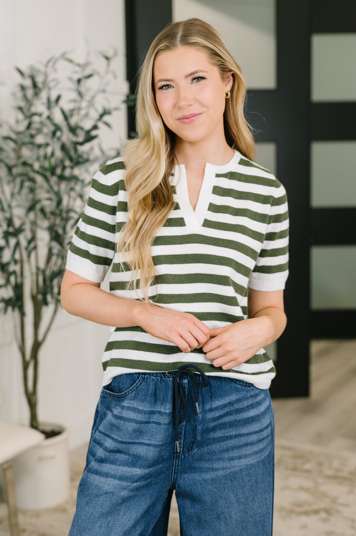 A woman wearing a green and white striped short-sleeve henley shirt with blue jeans, standing in a modern indoor setting with a potted plant and a dark-framed glass door in the background.