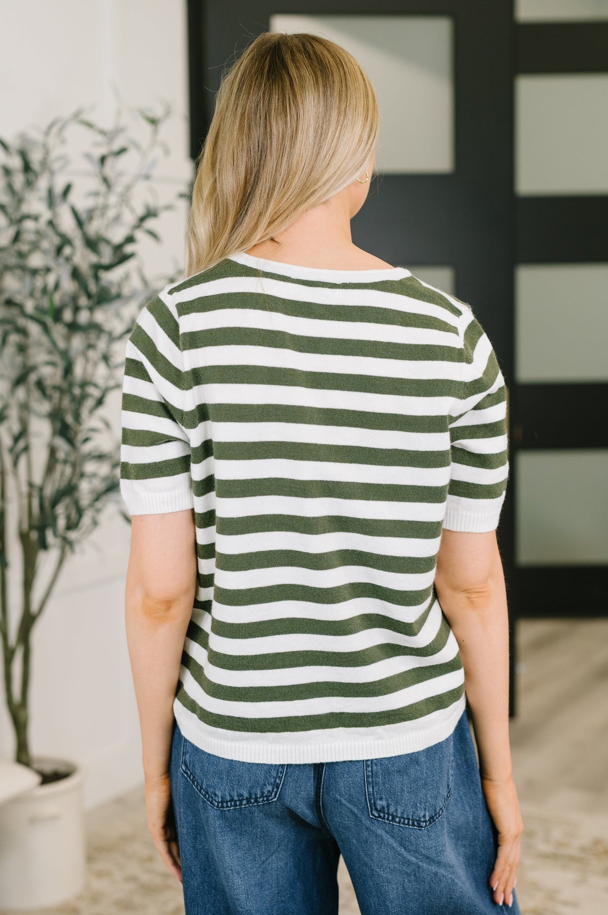 A woman viewed from behind wearing a short-sleeve knit top with horizontal green and white stripes, paired with blue jeans in an indoor setting.