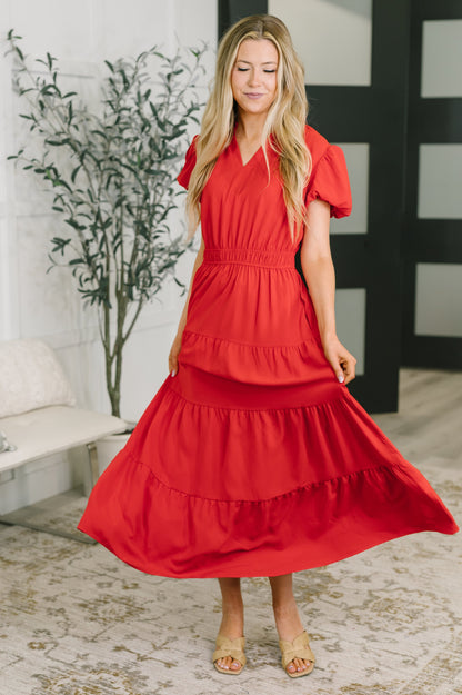 A woman wearing a bright red tiered midi dress with short puff sleeves and a V-neckline, standing in a bright indoor room with a patterned rug and a potted plant.