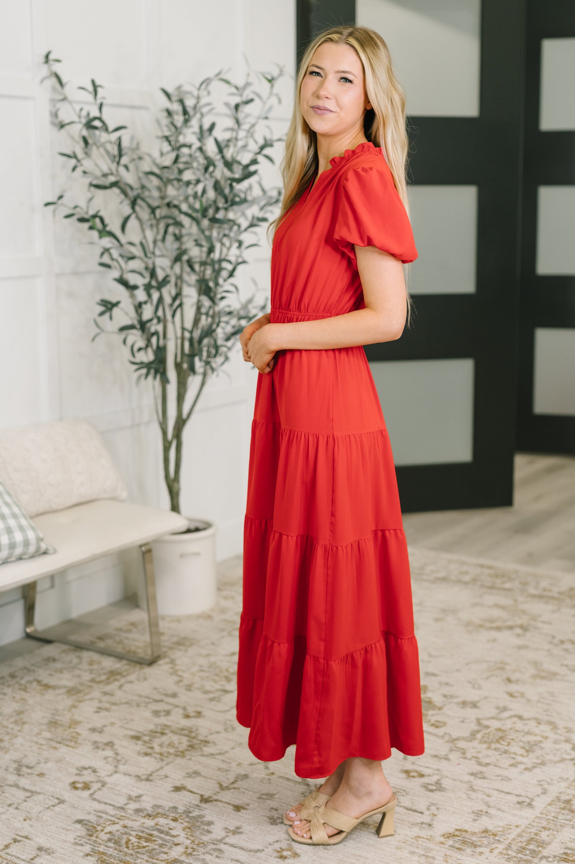 A woman modeling a bright red tiered maxi dress with short flutter sleeves, standing in a modern indoor setting with a patterned rug, potted plant, and white bench.