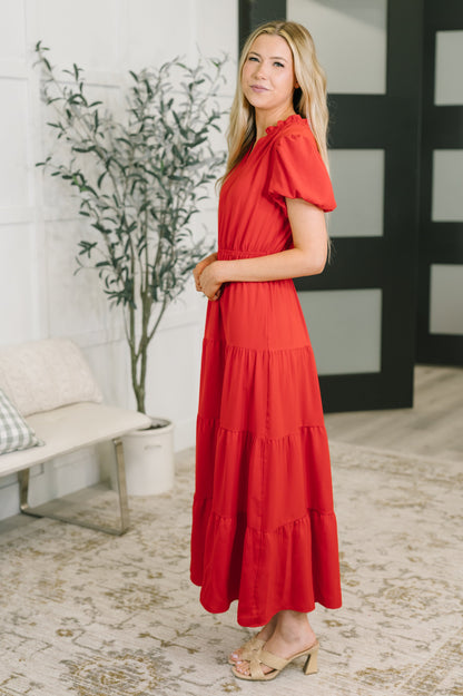 A woman modeling a bright red tiered maxi dress with short flutter sleeves, standing in a modern indoor setting with a patterned rug, potted plant, and white bench.