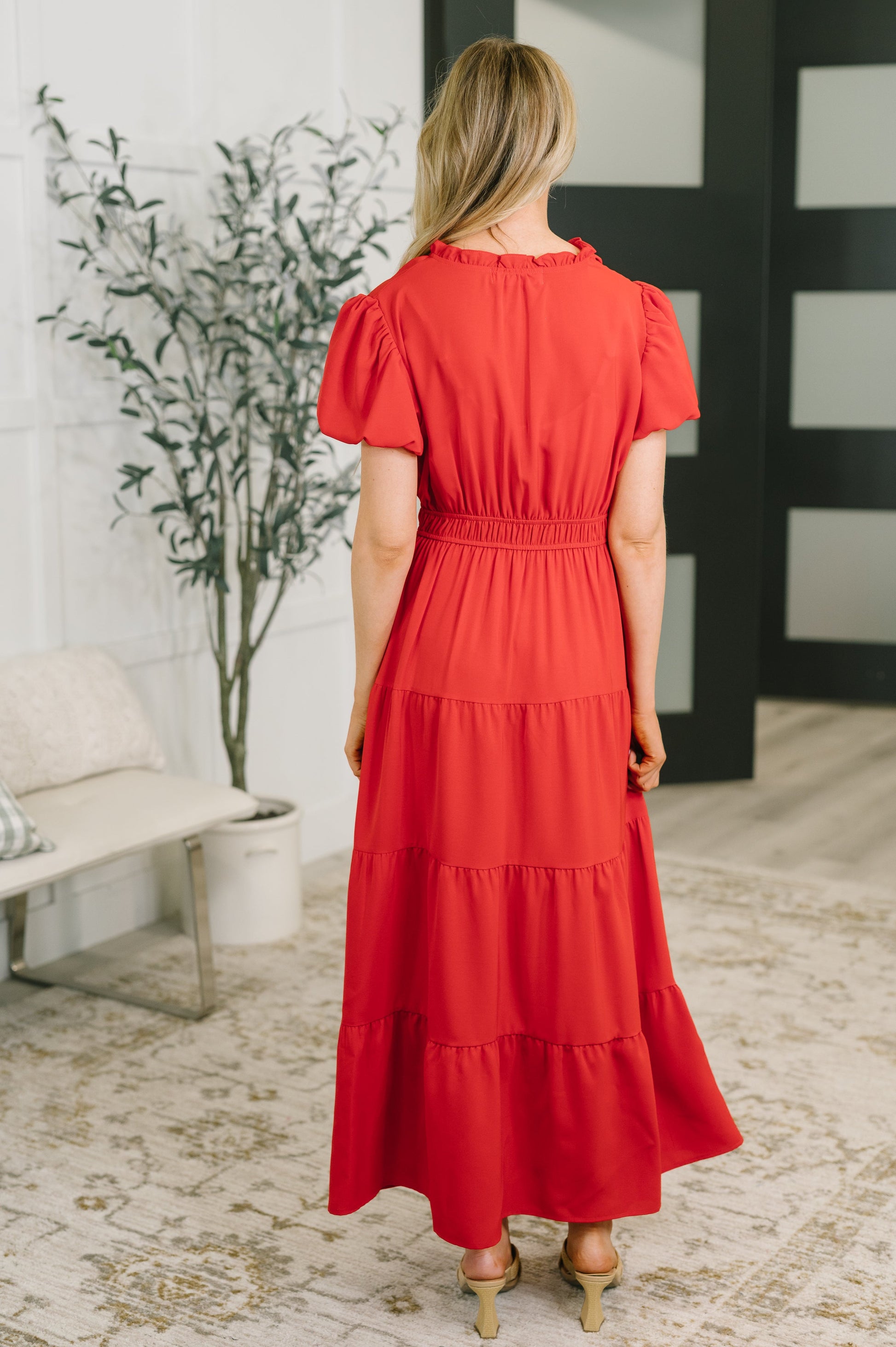 A woman viewed from behind wearing a bright red tiered midi dress with short puff sleeves and a smocked waist, standing in a modern indoor room with a bench, potted plant, and patterned rug.