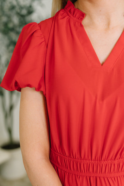 A close-up of a woman wearing a bright red dress with short puff sleeves, a ruffled neckline, and a gathered waist seam.