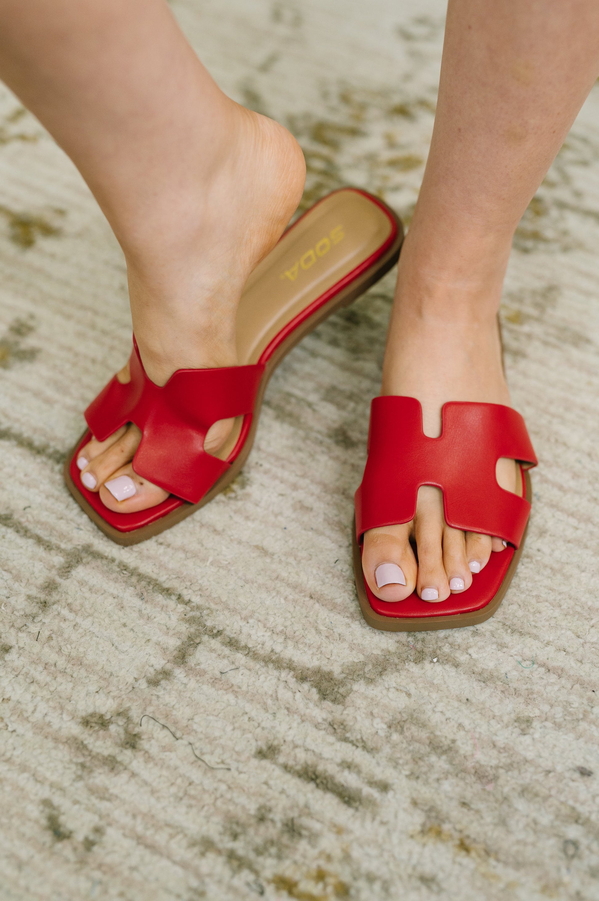 A person wearing red slide sandals with a cutout design and tan insoles, standing on a textured beige rug. The brand name 'soda' is visible on the insole.