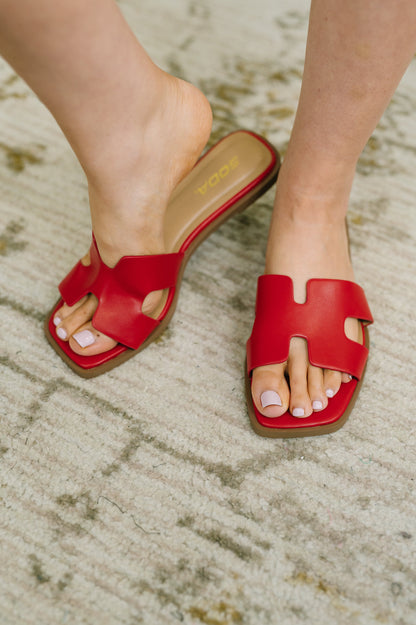 A person wearing red slide sandals with a cutout design and tan insoles, standing on a textured beige rug. The brand name 'soda' is visible on the insole.