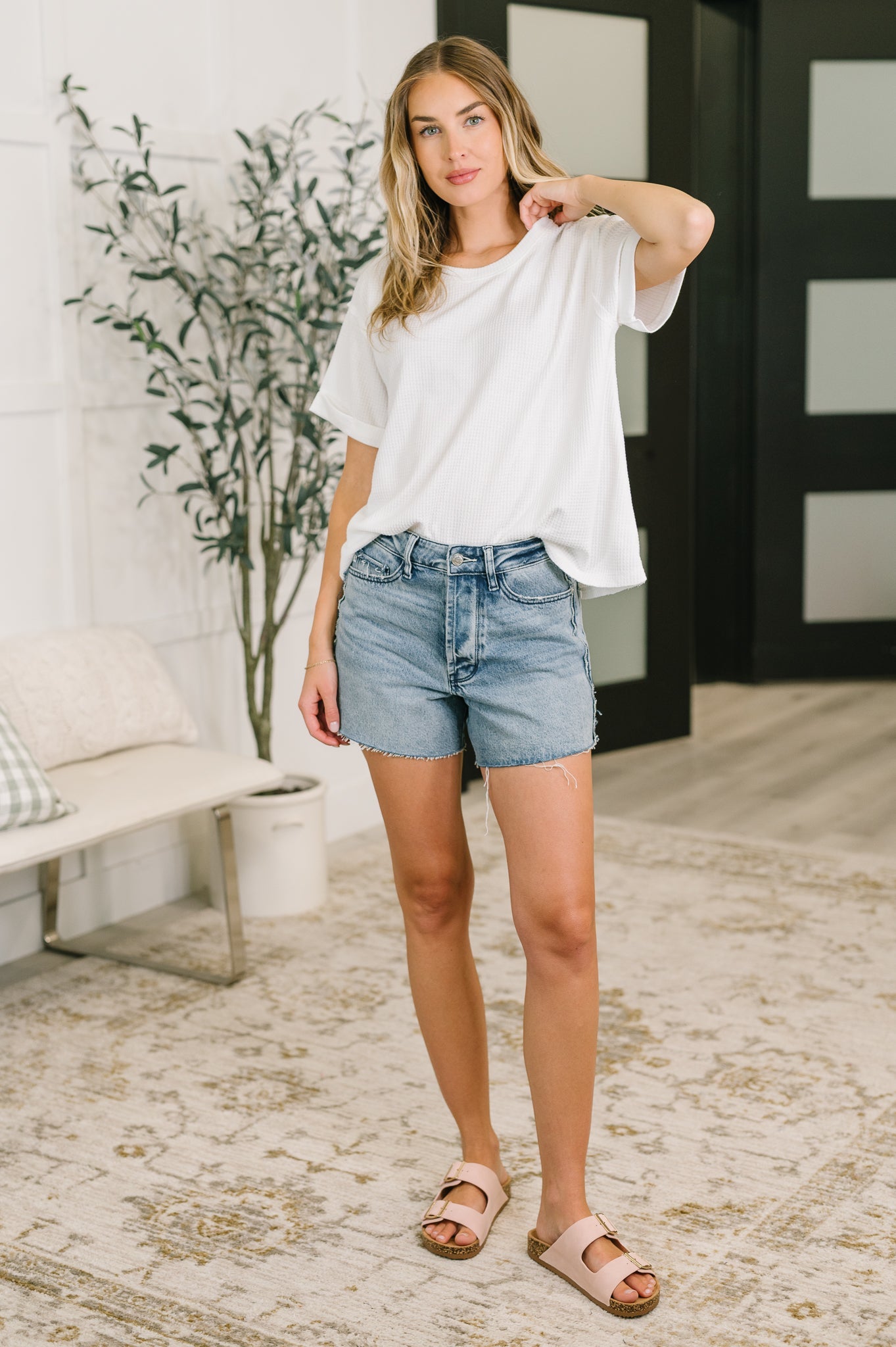 Woman wearing a white t-shirt and denim shorts standing in a room with a plant and furniture.