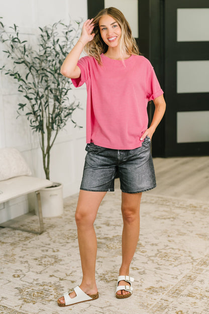 Woman wearing a pink top and denim shorts standing in a room with a plant and furniture.