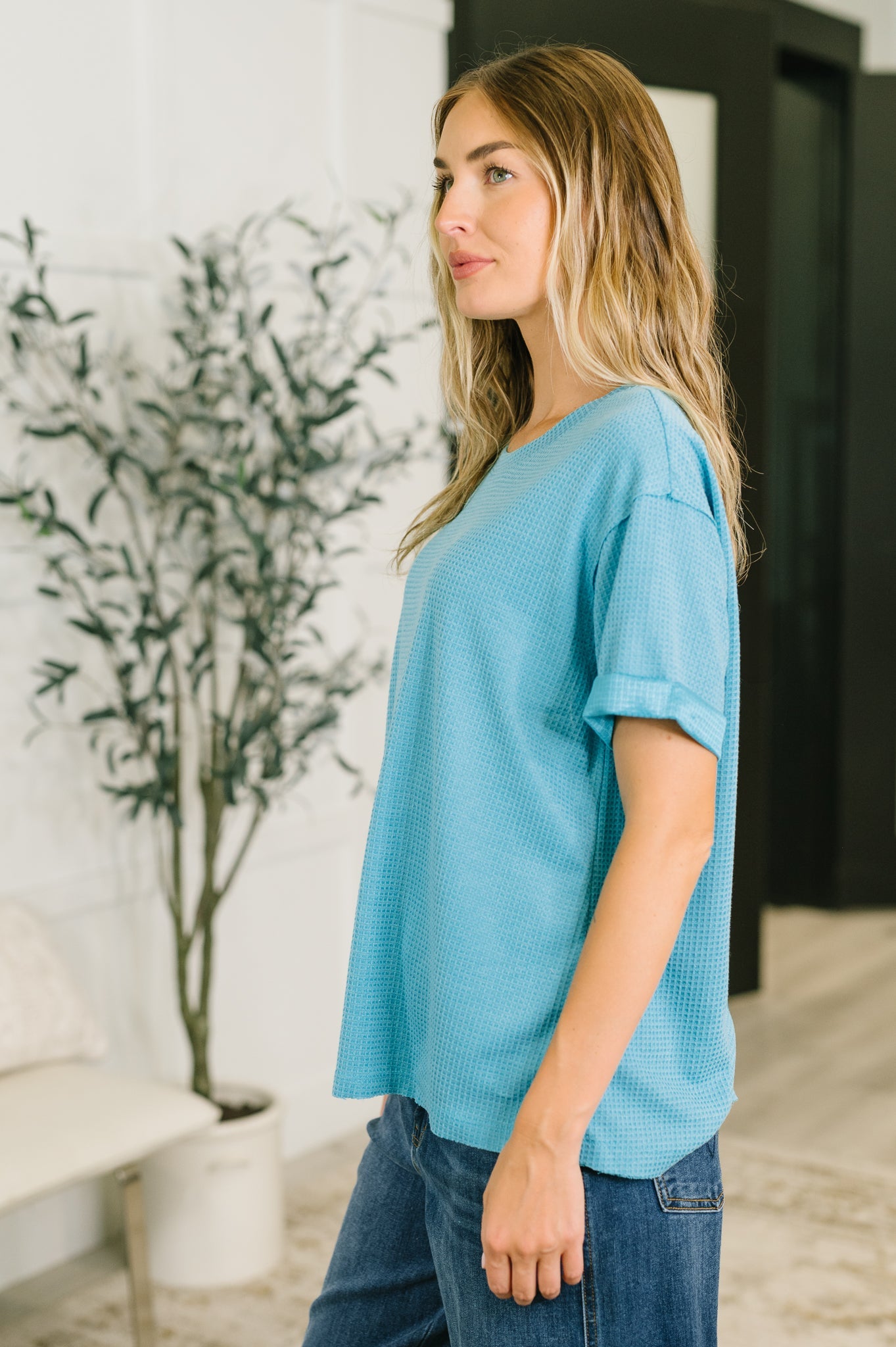 Woman wearing a blue top and jeans standing indoors with a plant in the background