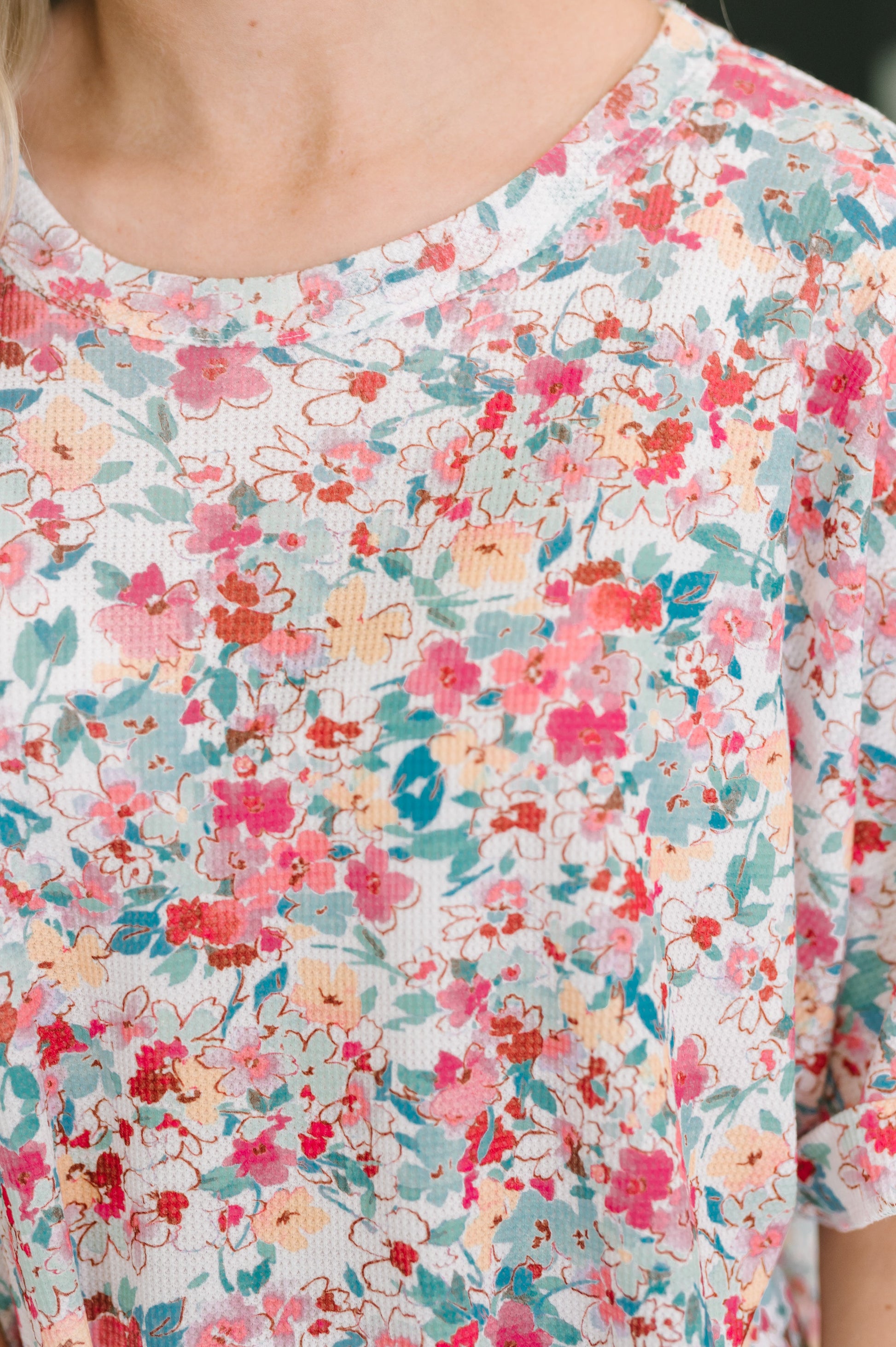 Close-up of a person wearing a short-sleeve top with a dense floral print in pink, coral, teal, and peach on a white base.